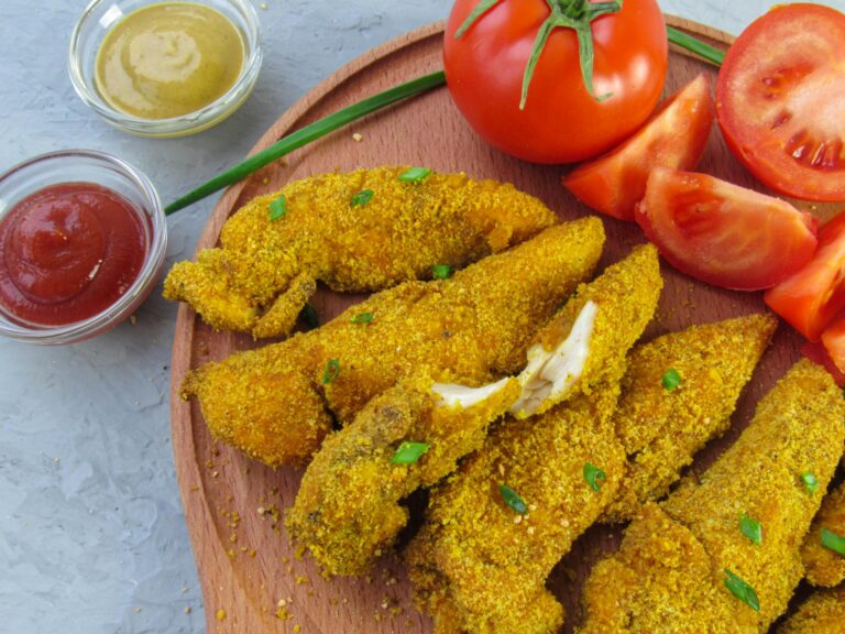 Crispy chicken fingers served with tomato slices and dipping sauces on a wooden tray.